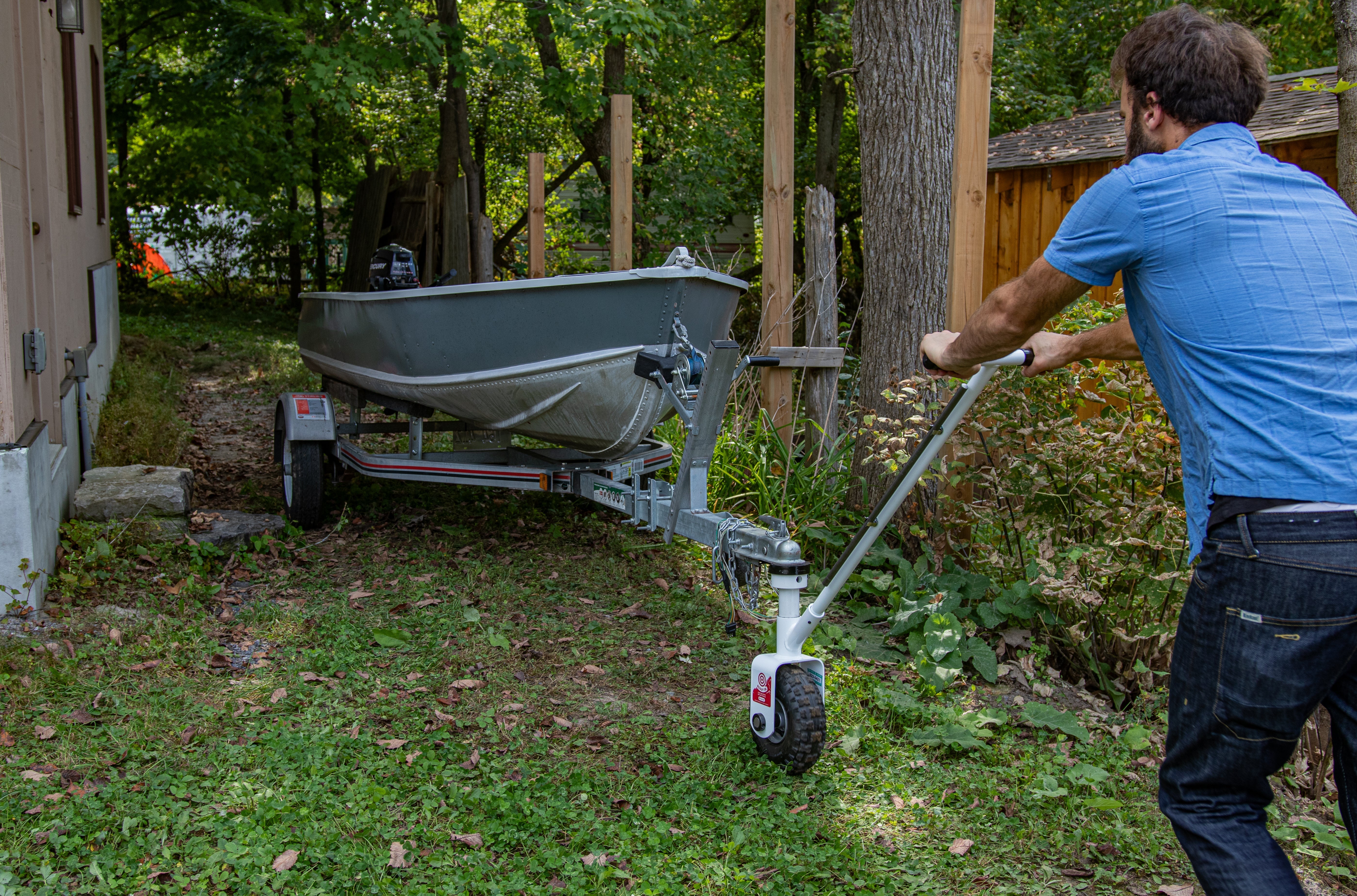 A man using a manual trailer dolly to pull a small boat trailer out of a rear yard.